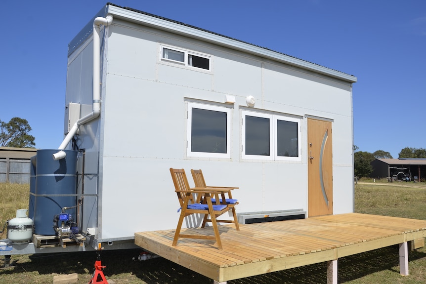 A tiny house with white exterior and  a timber deck wit two timber chairs on it.