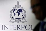 The silhouette of a man in a suit is visible in front of a wall with the Interpol logo on it.