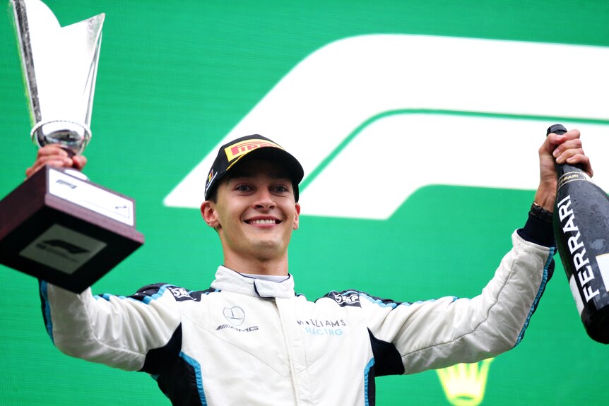 George Russell smiles while holding a trophy in one hand and a large bottle of champagne in the other