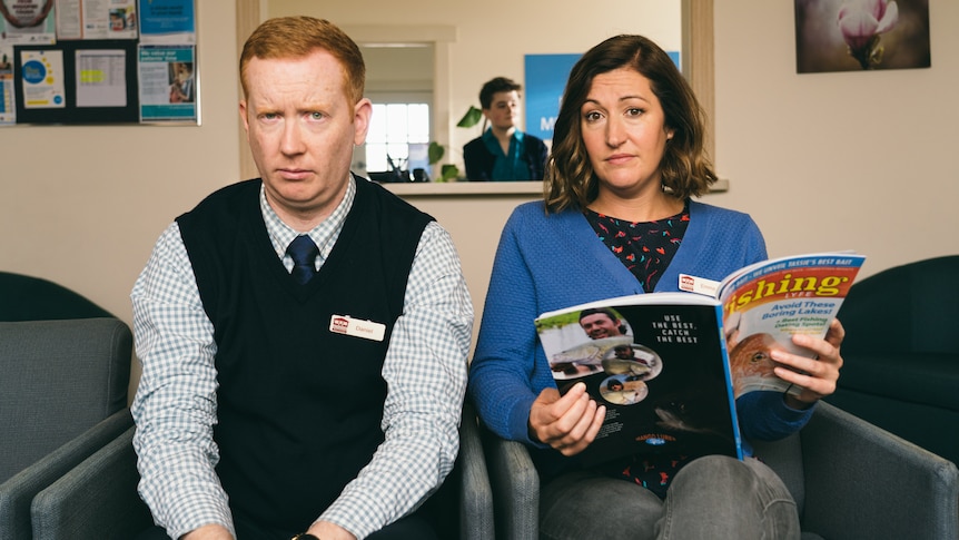 Luke and Celia sit in a country health centre waiting room, in their Rosehaven costumes, looking concerned at the camera.