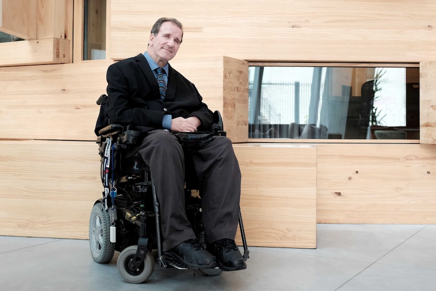 Simon Darcy, in a wheelchair, near wooden wall