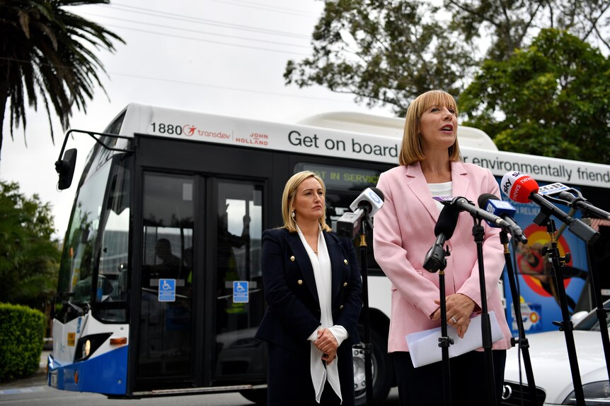 Two women in blazers stand in front of a large bus facing a line of microphones