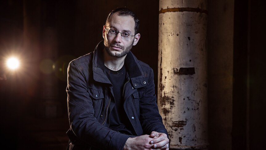 Man wearing glasses seated with hands together on knee in darkly lit industrial space and light shining from behind.