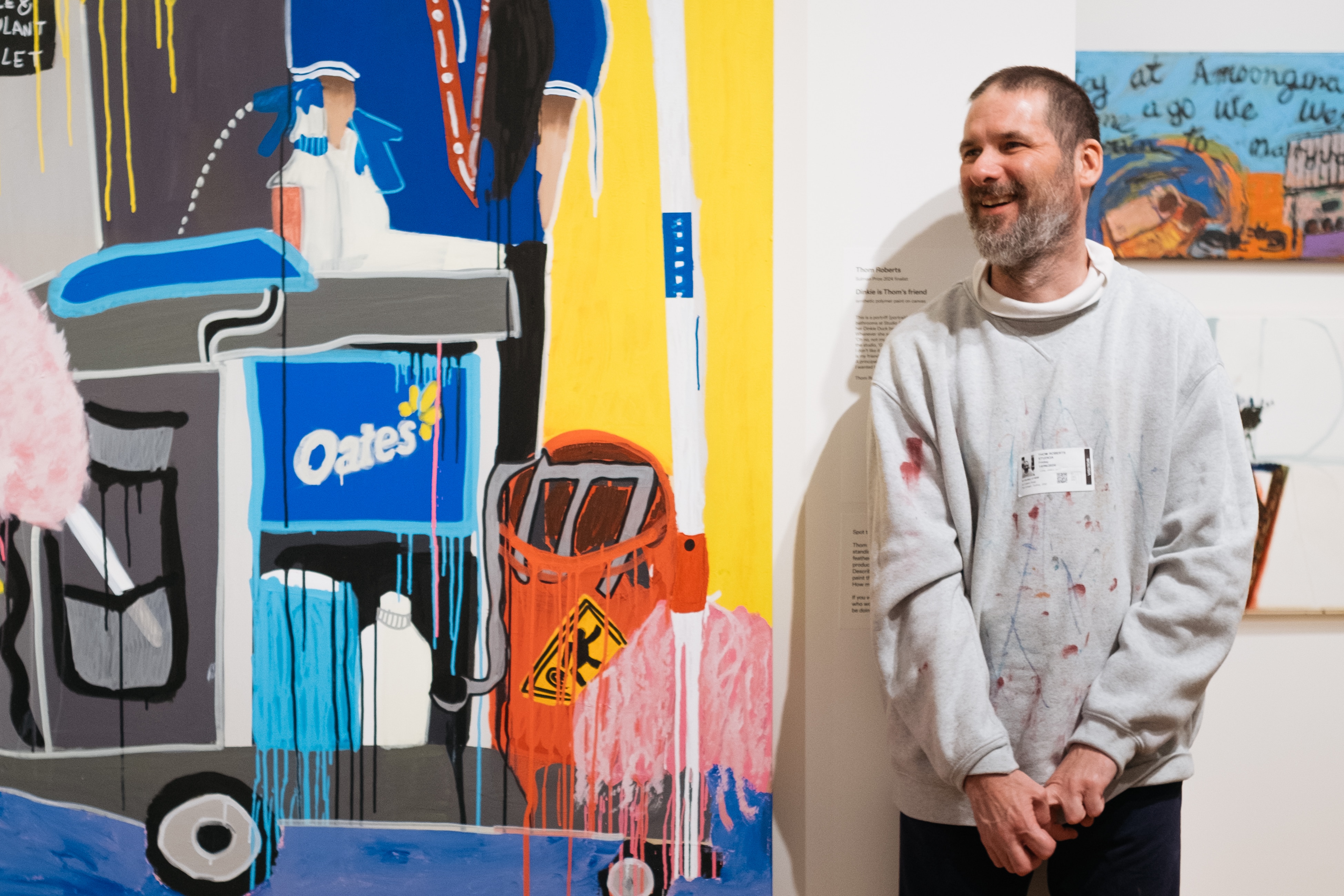 The artist wears a grey jumper covered in paint, and has save brown and grey hair. He stands smiling in AGNSW next to paintings