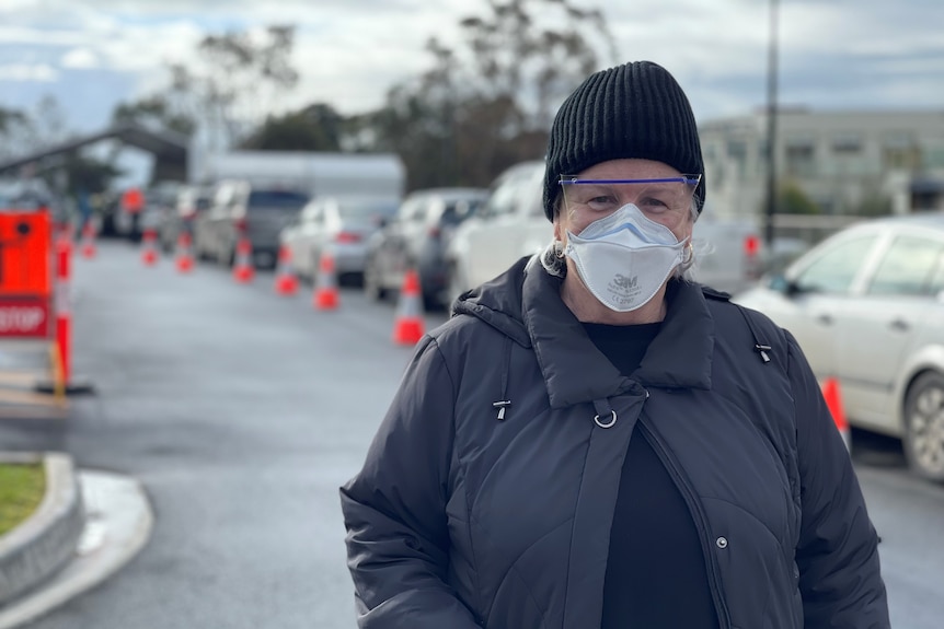 A woman wearing a mask stands in front of a line of cars.
