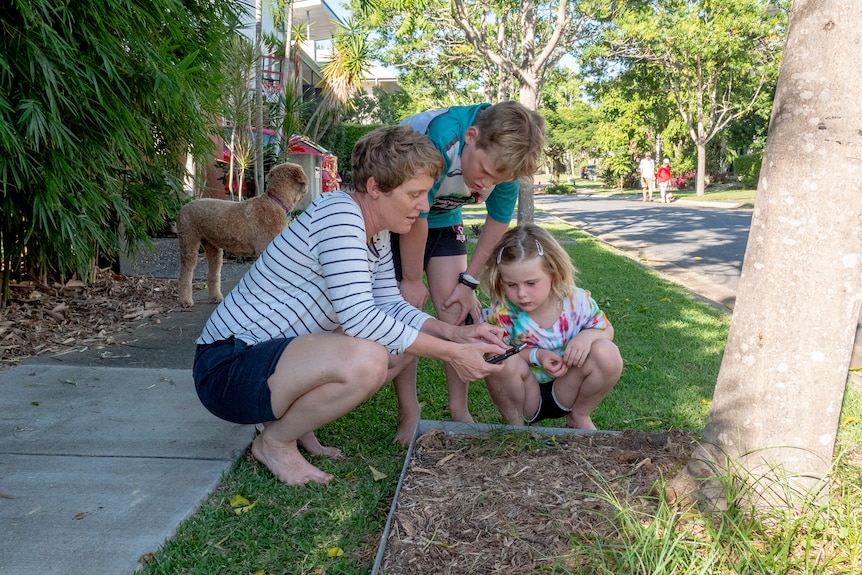 A woman and her two young children couch on the nature strip, looking at an app on her phone.