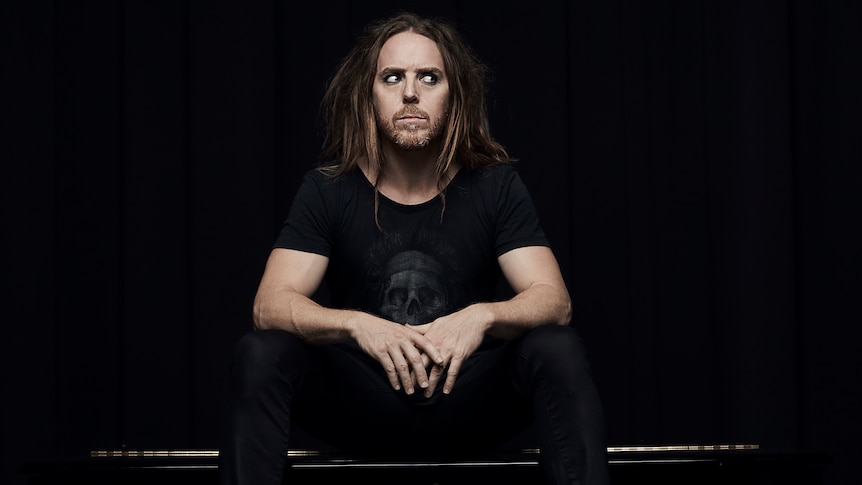 A man with long straight hair sits barefoot on a black grand piano.