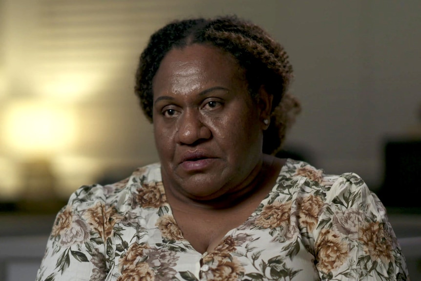 Sarah Kiniafa wearing a floral dress sits in a darkened room with a lamp glowing in the background.