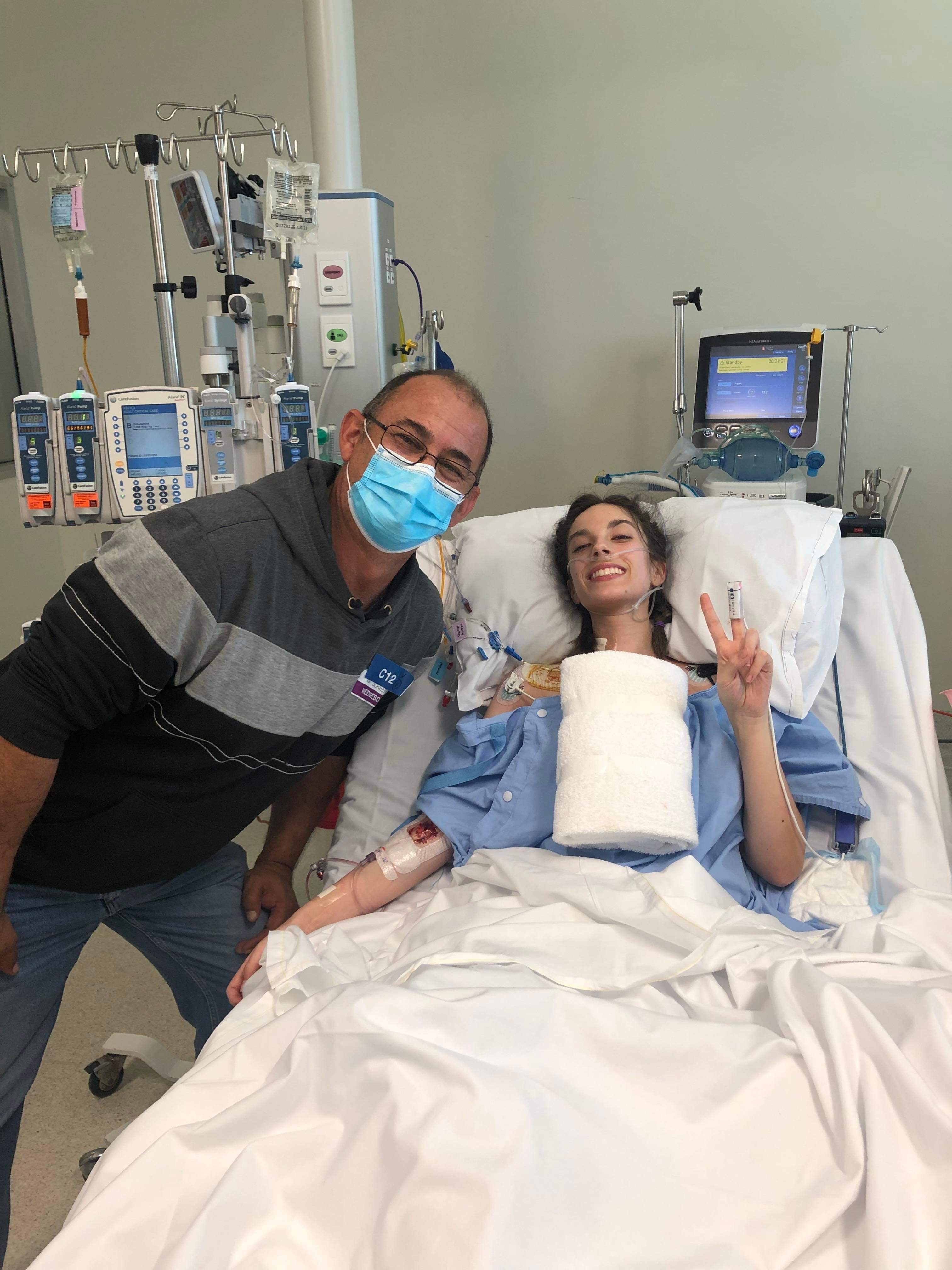 A young smiling woman in a hospital bed, with an older man in a surgical mask standing next to her