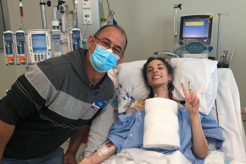 A young smiling woman in a hospital bed, with an older man in a surgical mask standing next to her