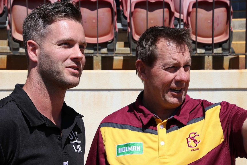 Jake Edwards stands on the left next to Peter Capes in front of stadium seating as they both look off to the right.