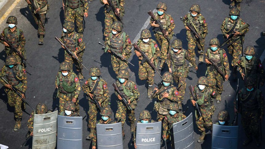 Armed soldiers in fatigues and face masks walk along a street with rifles pointed at the ground.
