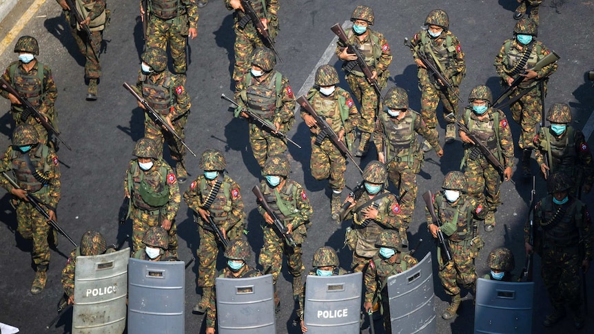 Armed soldiers in fatigues and face masks walk along a street with rifles pointed at the ground.