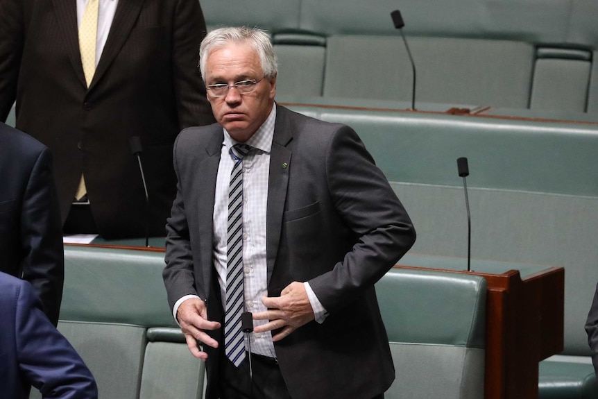 Drum is standing, looking across the chamber. He looks concerned.