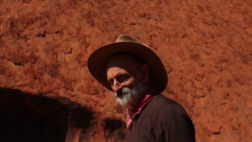 An older man in an akubra stands in front of a big red rock in the dessert.