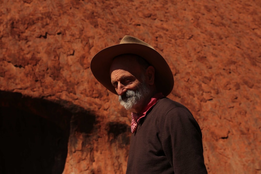 An older man in an akubra stands in front of a big red rock in the dessert.