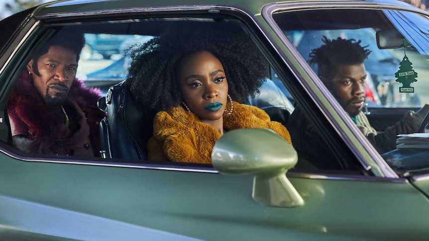 Still from They Cloned Tyrone: Jamie sits in the back seat of a mint old-fashioned car with Teyonah front right and John left.