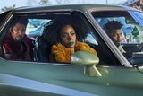 Still from They Cloned Tyrone: Jamie sits in the back seat of a mint old-fashioned car with Teyonah front right and John left.