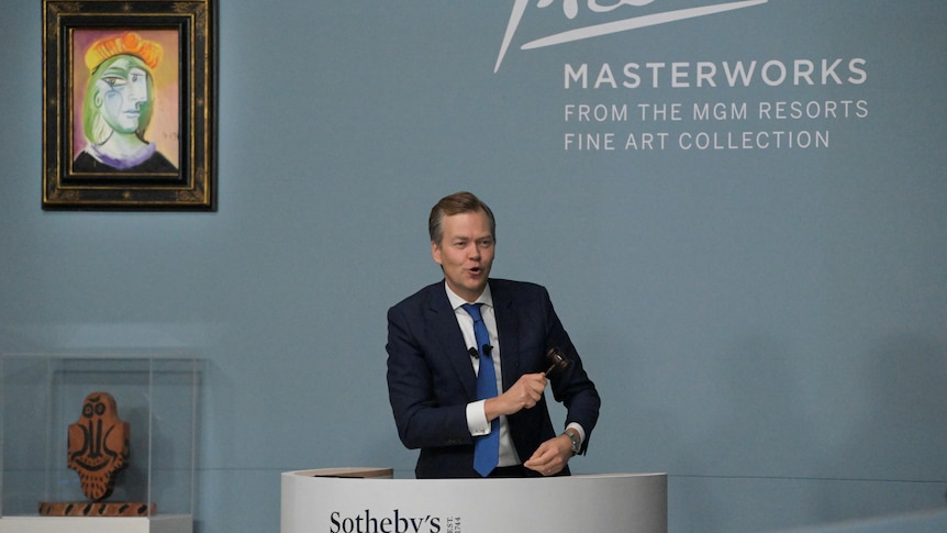 An auctioneer holding a gavel stands in front of a painting. 