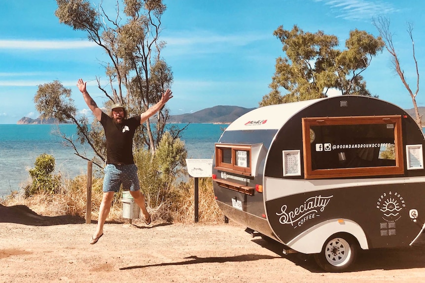 A man leaps into the air next to a vintage-looking caravan, with the ocean in the background.