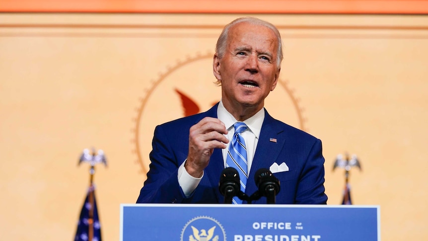 A man with white hair stands a podium saying Office of the President Elect wearing a blue suit and tie.