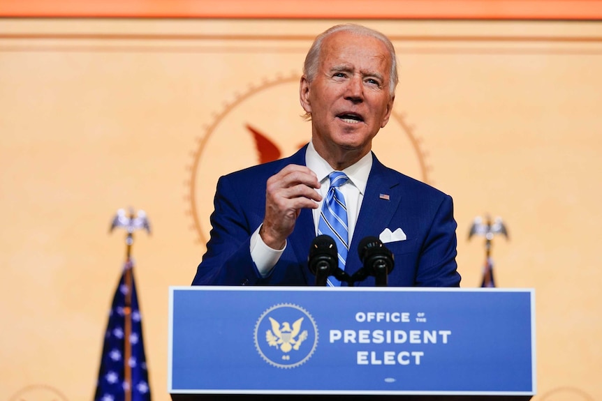 A man with white hair stands a podium saying Office of the President Elect wearing a blue suit and tie.