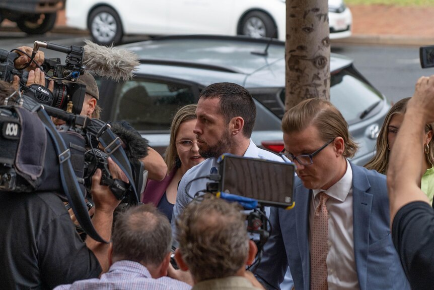 A man in a blue dress shirt is surrounded by media and camera operators. 