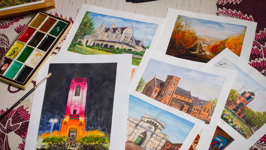Artworks in watercolour mainly of buildings lying on a table with paints and a brush nearby