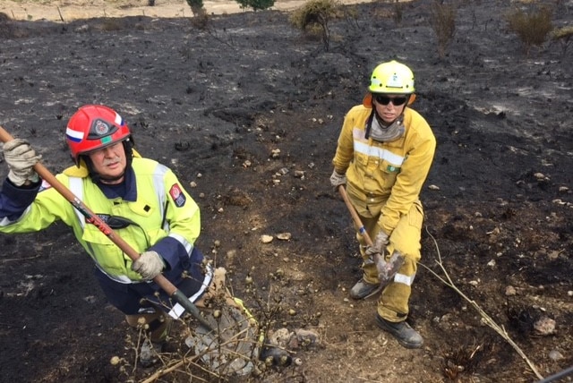 Two firefighters hold a pick axe and a shovel in a burnout field.