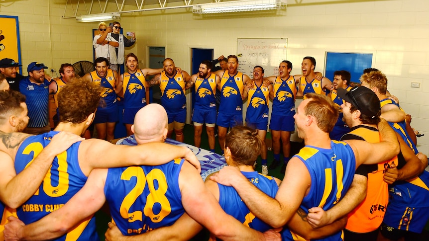 A group of men wearing blue and yellow football jumpers stand in a circle singing. 