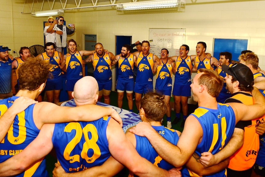 A group of men wearing blue and yellow football jumpers stand in a circle singing. 