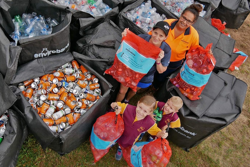 Two women and two children stand in front of a large collection of bottles and cans collected in bales for recycling