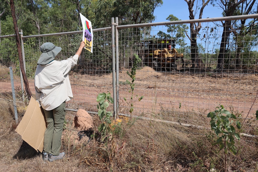 A person holds a banner up near a fence while a bulldozer clears land.
