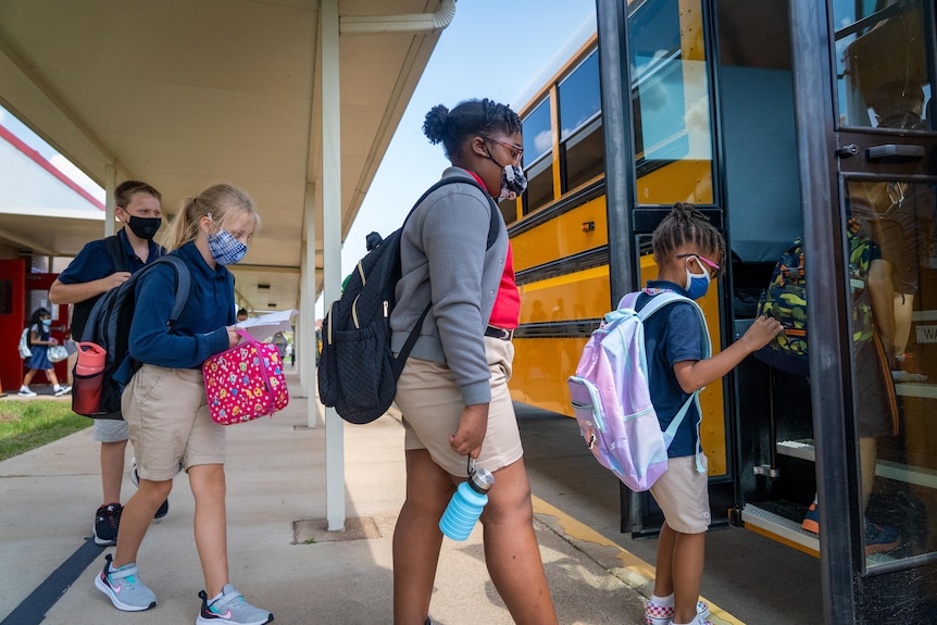 Children in face masks line up to board a big yellow school bus 