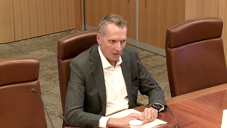 Dr Paul Scott wears a grey suit and a white open-necked shirt and sits at a table in front of a microphone