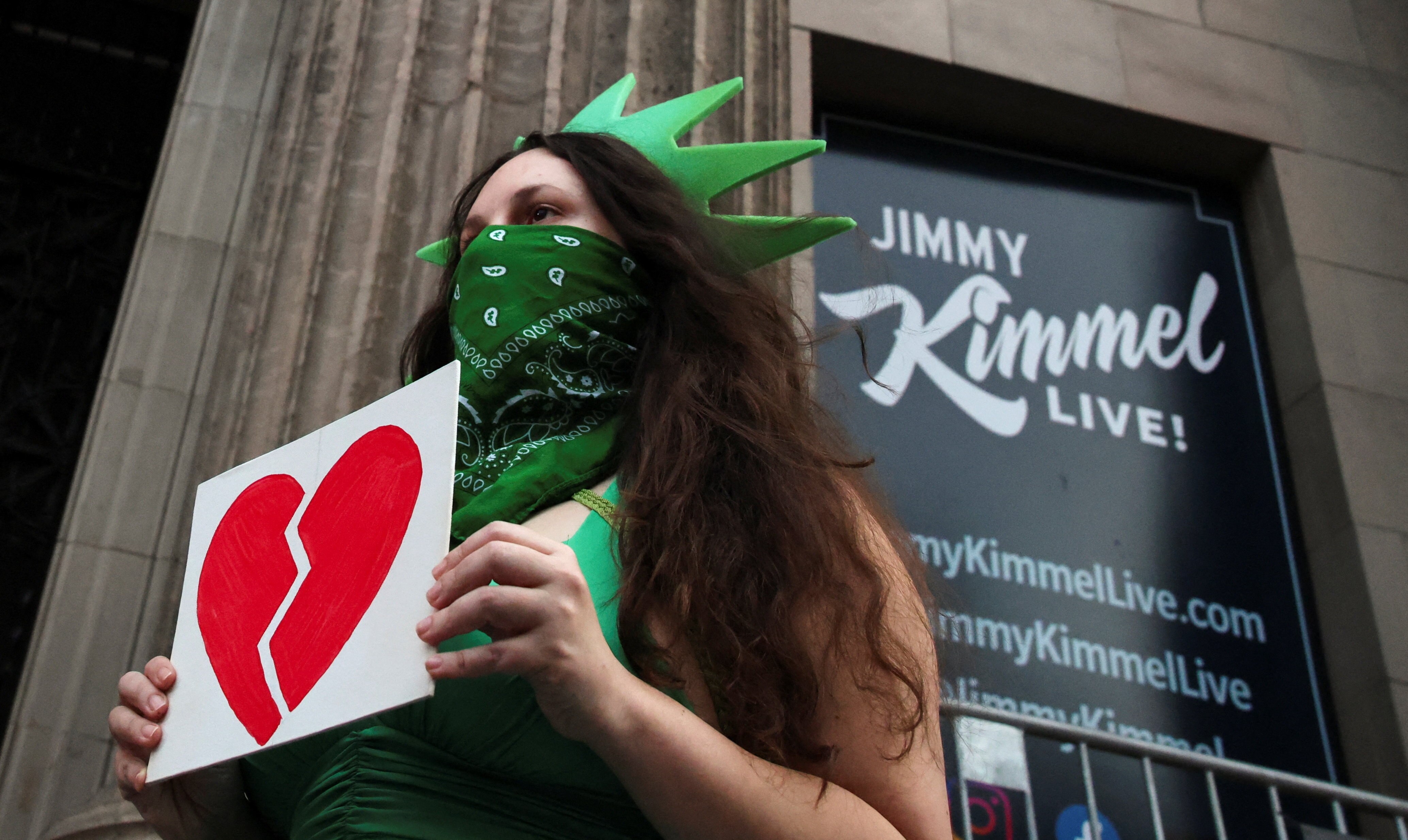 A woman wearing a Statue of Liberty outfit and mask, holding a sign with a broken heart.