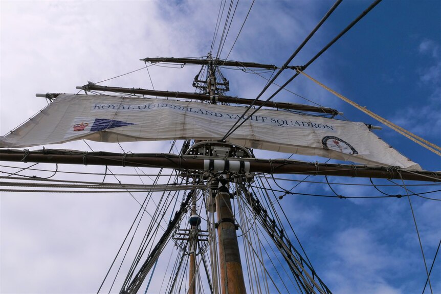 Looking up at a mast of a ship, from the deck, with a sail being released 