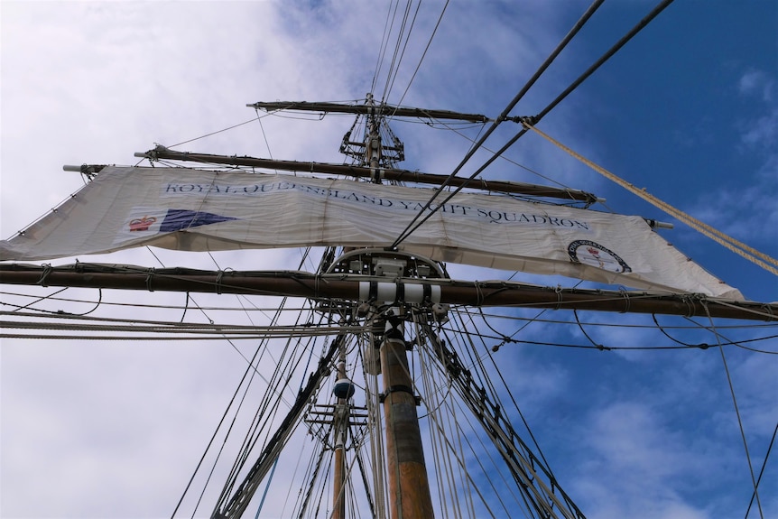 Looking up at a mast of a ship, from the deck, with a sail being released 