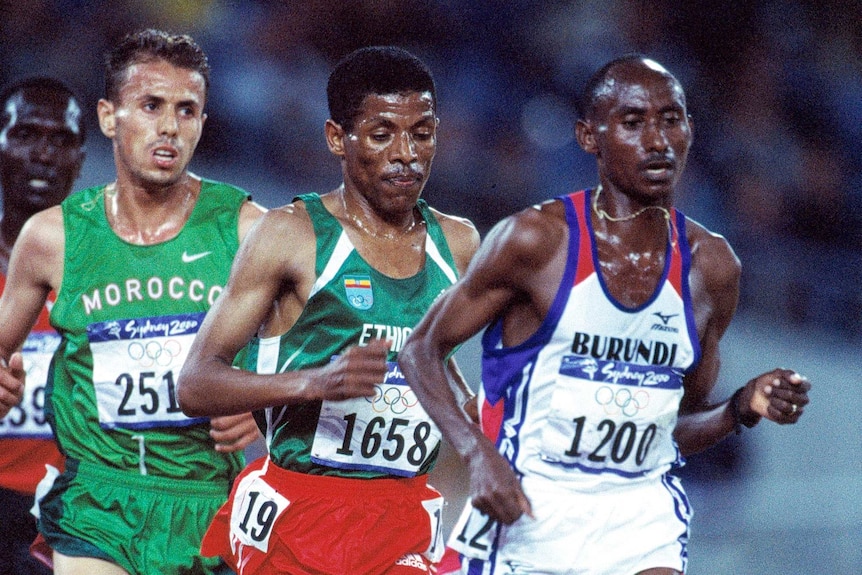 Aloys Nizigama runs in a white vest in front of Haile Gebrselassie in a green vest
