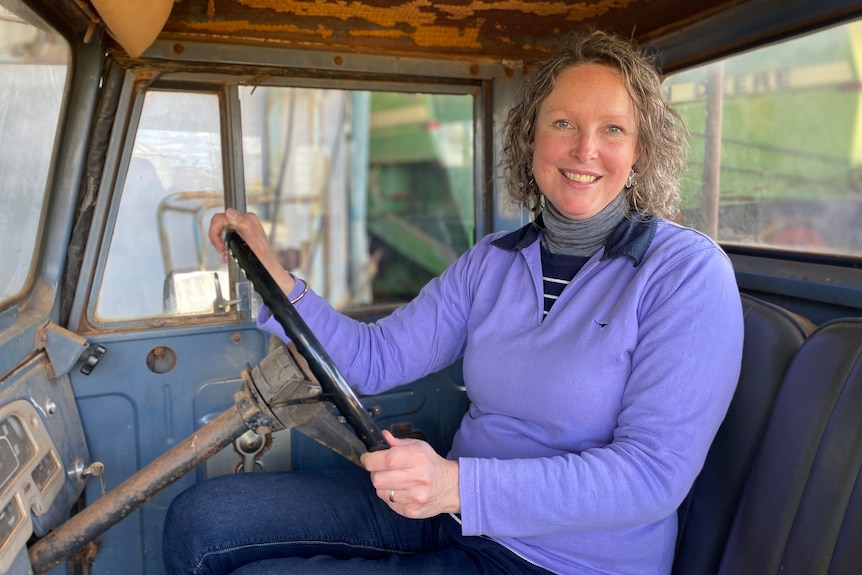 Peri McIntosh, a woman with curly grey hair wearing a lilac jumper, sits in the cab of an old car amid tractors.