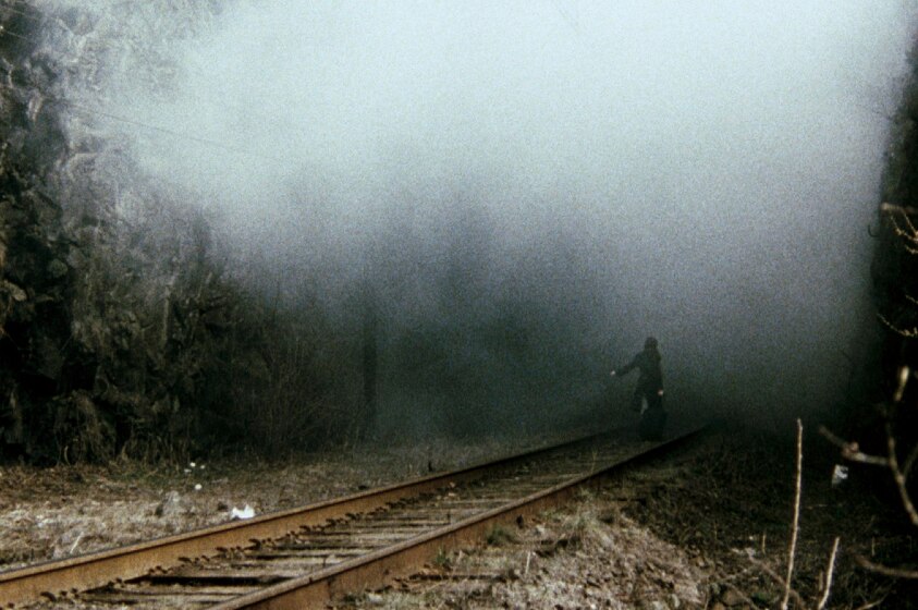 A human figure in long black coat stands on rural train track thrusting body towards oncoming large fog or mist.