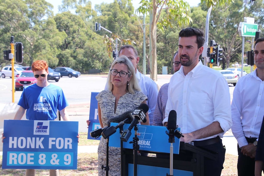 A wide shot of Zak Kirkup speaking at a media conference outdoors with colleagues and a man holding a sign behind him.