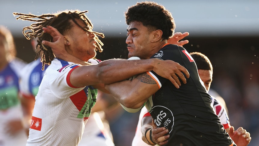 A Wests Tigers NRL player carries the ball as he is tackled by a Newcastle Knights opponent.