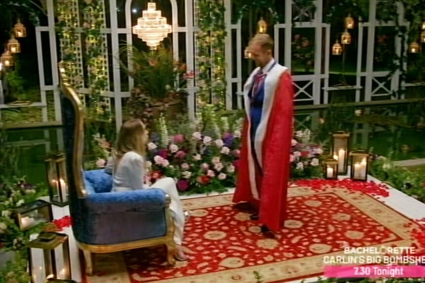 Jess Glasgow dressed in red regal robes standing in front of The Bachelorette Angie Kent sitting in a blue throne.