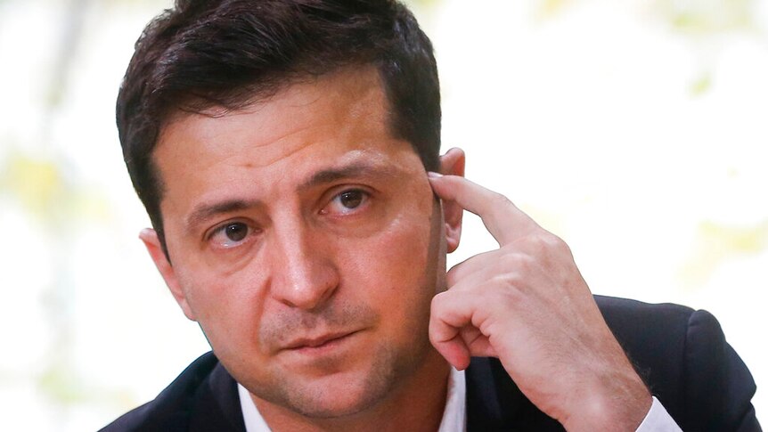 Volodymyr Zelenskiy listens to a question and scratches the side of his face. His expression is serious.