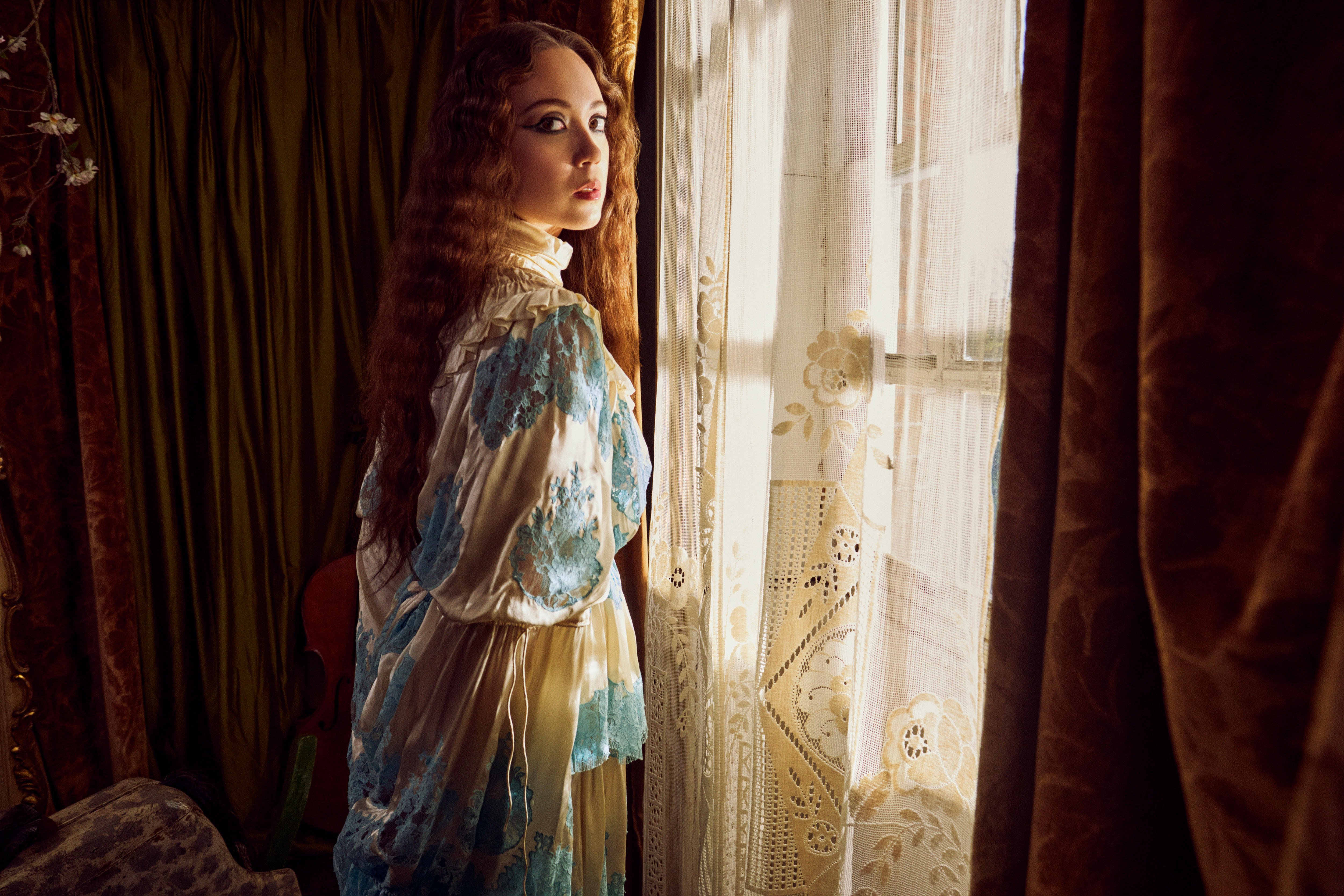 Laufey looks over her shoulder at the camera as she stands at a window with a lace curtain