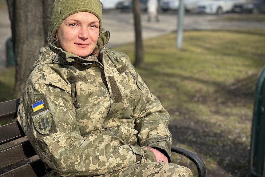 A woman wearing army fatigues sits on a park bench and smiles.