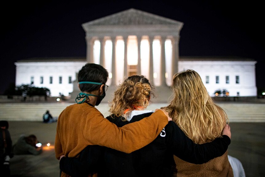Three women with their arms around each other stand in front of the US Supreme Court
