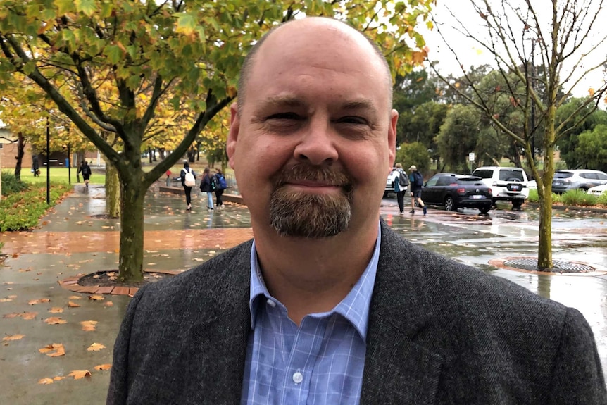 Head and shoulders shot of a bald, middle-aged man dressed in bookish attire. The weather is overcast.