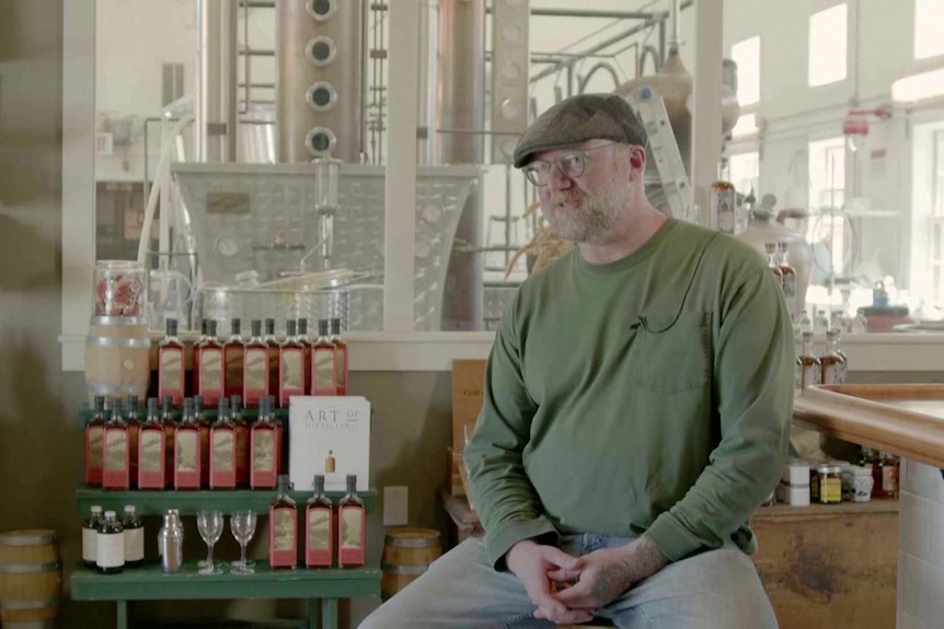 een man met een pet en een bril zit in een distilleerderij naast flessen whisky